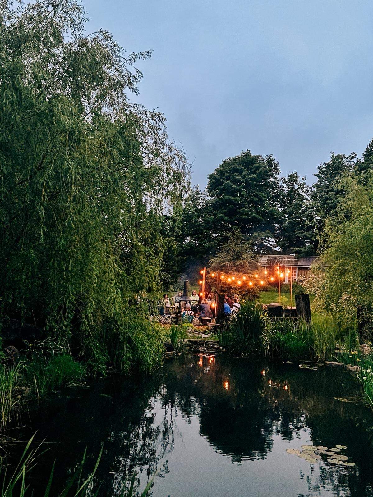 A gathering lit by string lights at dusk, reflected in a still pond framed by willow branches.