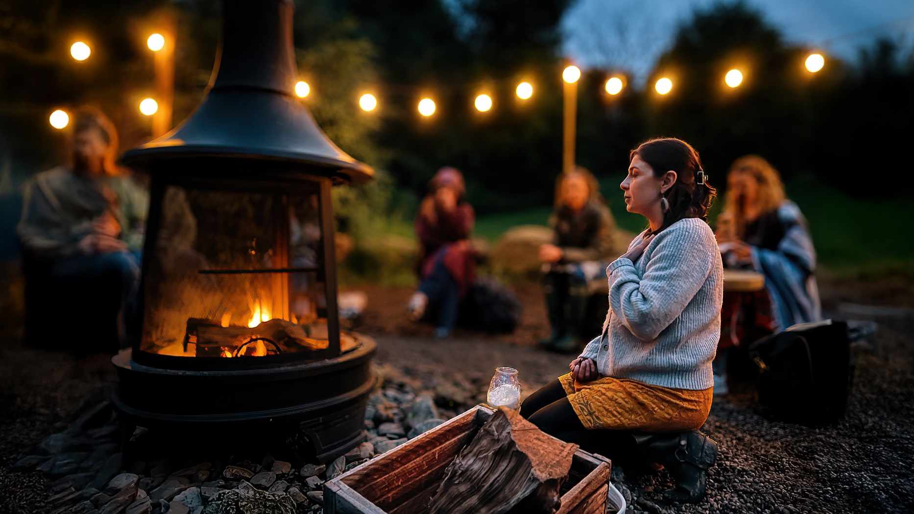 Sydney Blum sitting beside a lit outdoor fire at dusk, with other women gathered under strung lights behind her.