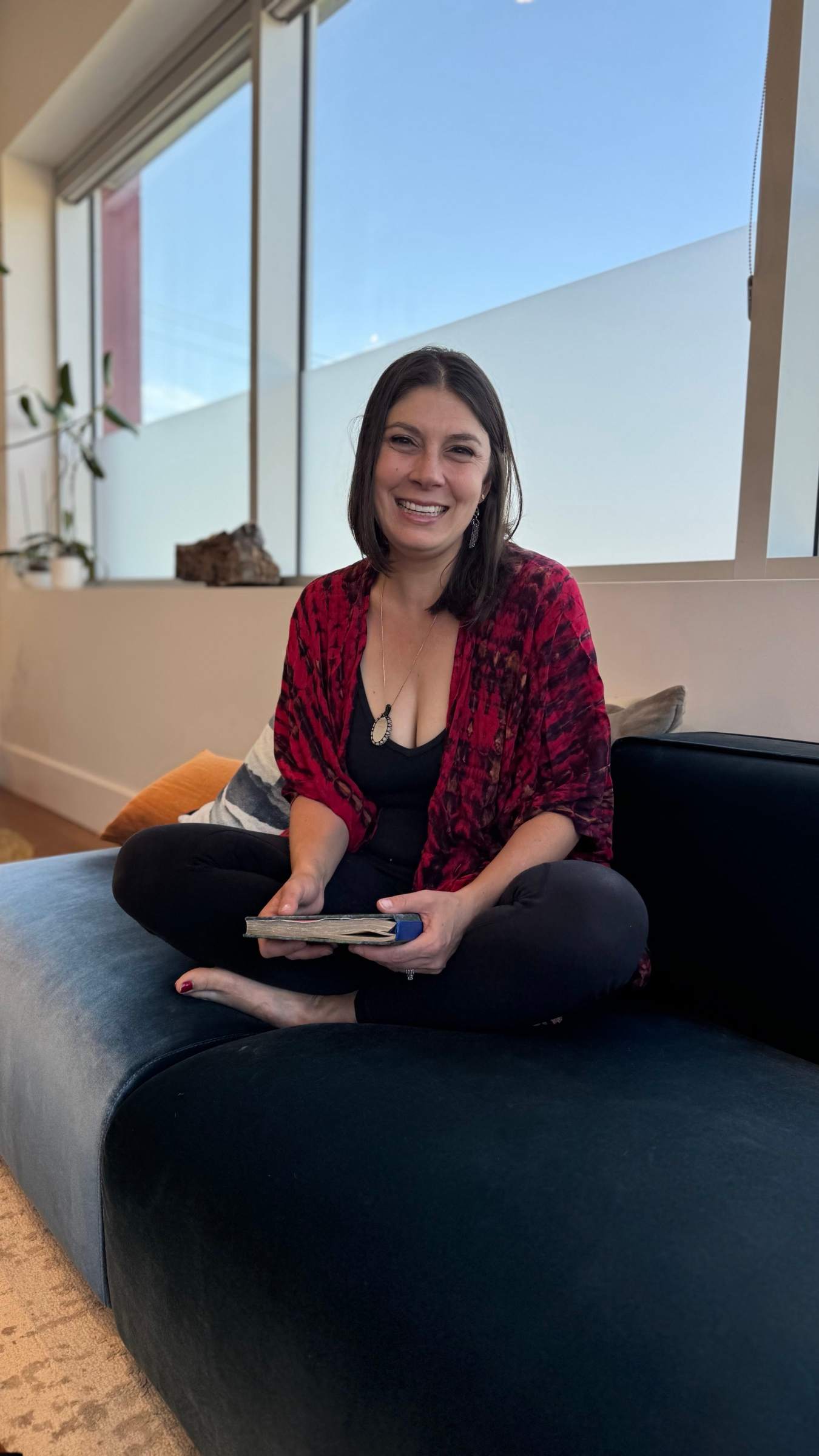 Sydney Blum sitting cross-legged on a couch in a red tie-dye wrap, holding a journal, smiling.